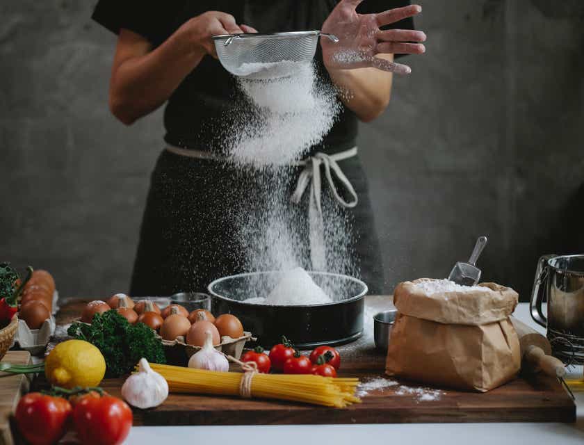 A person sifting flour at a table laden with culinary products.