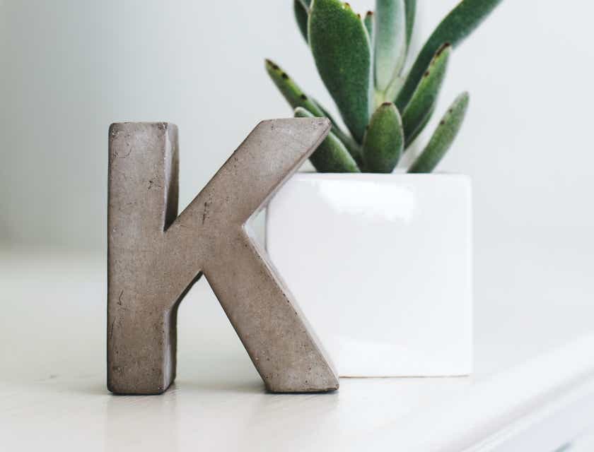 Hiasan huruf K berdiri di atas meja di sebelah pot tanaman.