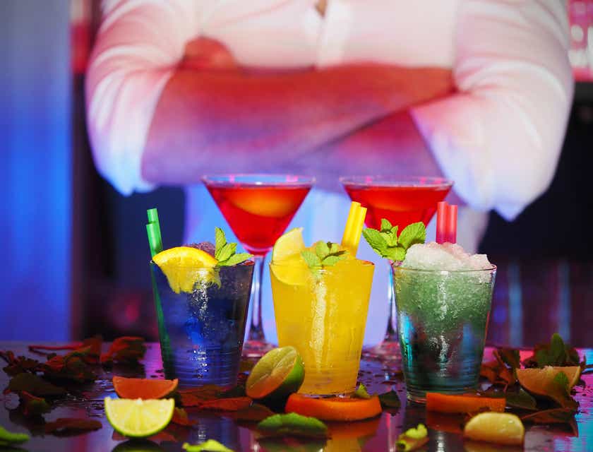 Een barman met gekruiste armen achter verschillende, felgekleurde cocktails.