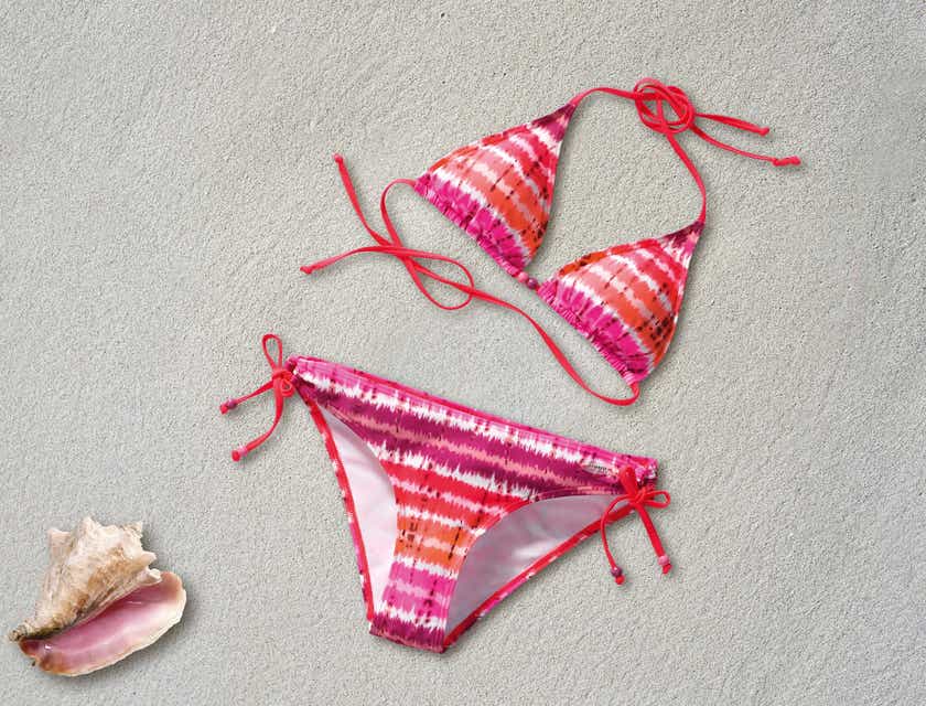 A pink and orange tie-dye bikini laid out on soft sand beside a sea shell.