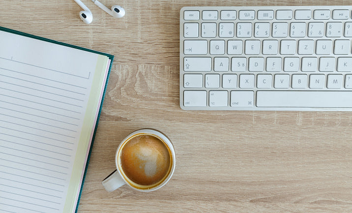 A keyboard, notebook, and earbuds lying on a table near a cup of coffee, ready to be used to write a blog.