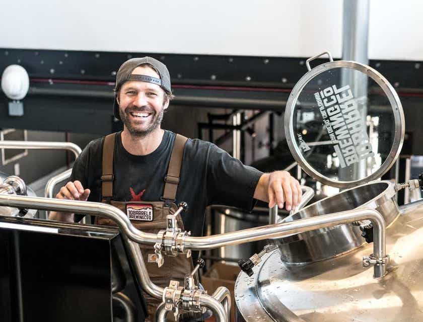Ein lächelnder Mann steht in einer Brauerei neben Metallschläuchen und einem großen Stahlfaß.