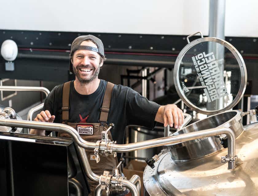 Seorang pria berdiri dan tersenyum di belakang fermentor sebuah brewery.
