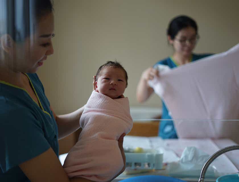 Eine Doula hält stolz ein in eine rosa Decke eingewickeltes neugeborenes Baby in ihren Armen.