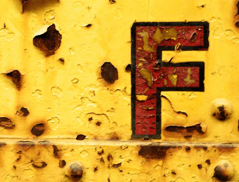 Rdzewiejąca litera F na tle ściany z odchodzącą, żółtą farbą.