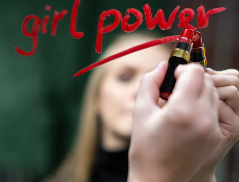 Una mujer usando un lápiz labial para escribir una popular frase de empoderamiento femenino en un espejo en un logo para empoderamiento femenino.