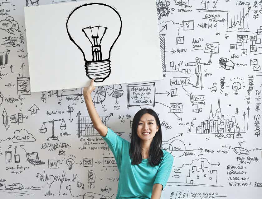 Seorang wanita duduk di depan papan tulis dan memegang gambar bola lampu yang inovatif.