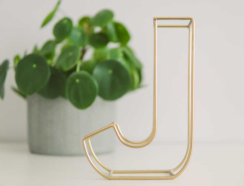 Złote logo z literą „J” w formie metalowej dekoracji na tle białej ściany i rośliny.
