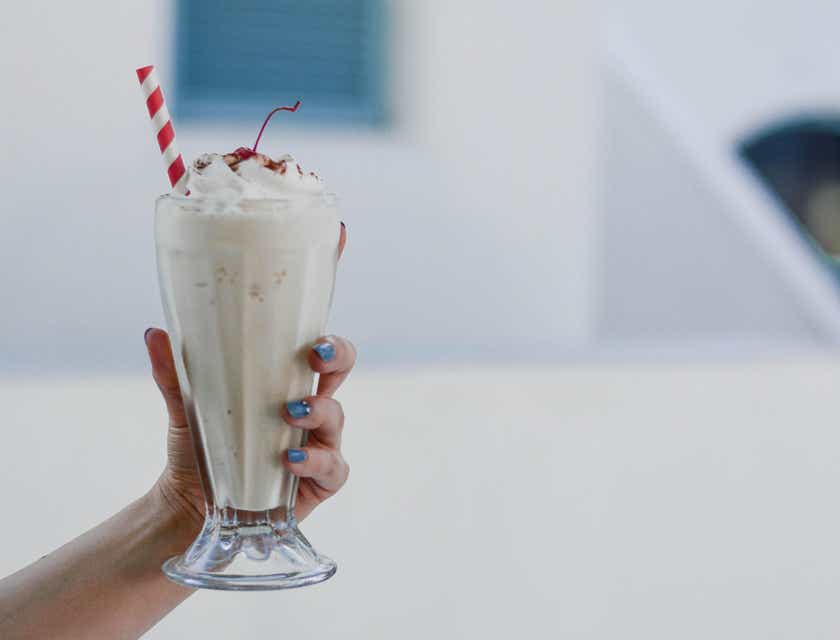 Sebuah tangan memegang milkshake rasa vanilla dalam gelas transparan.
