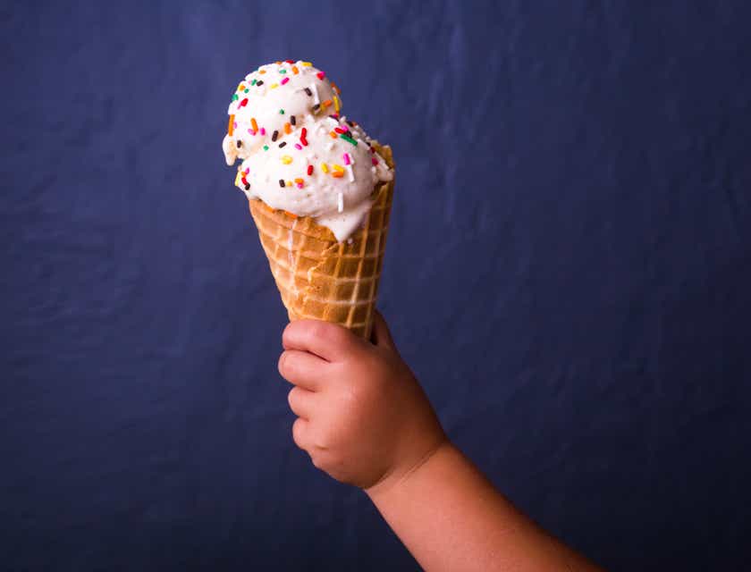 La mano de un niño sosteniendo un cono con una bola de nieve de fresa con chispas de colores con un fondo azul oscuro en una tienda de helados.