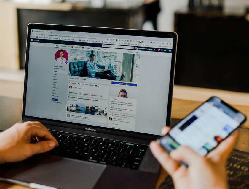 Una persona que sostiene un teléfono inteligente y se sienta frente a una computadora portátil abierta que muestra una página de redes sociales en un logo para empresas de medios.