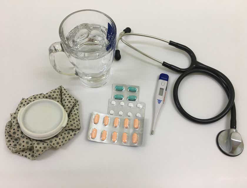 Przyrządy medyczne – stetoskop, termometr oraz leki – oraz szklanka wody na białym stole.