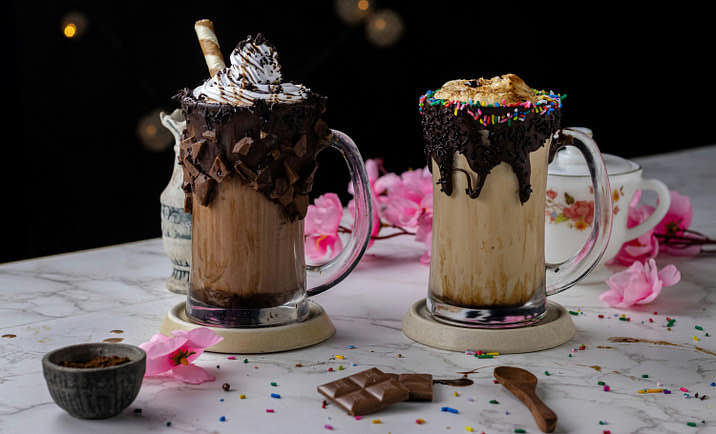 Two elaborate chocolate milkshakes sitting on a messy table.