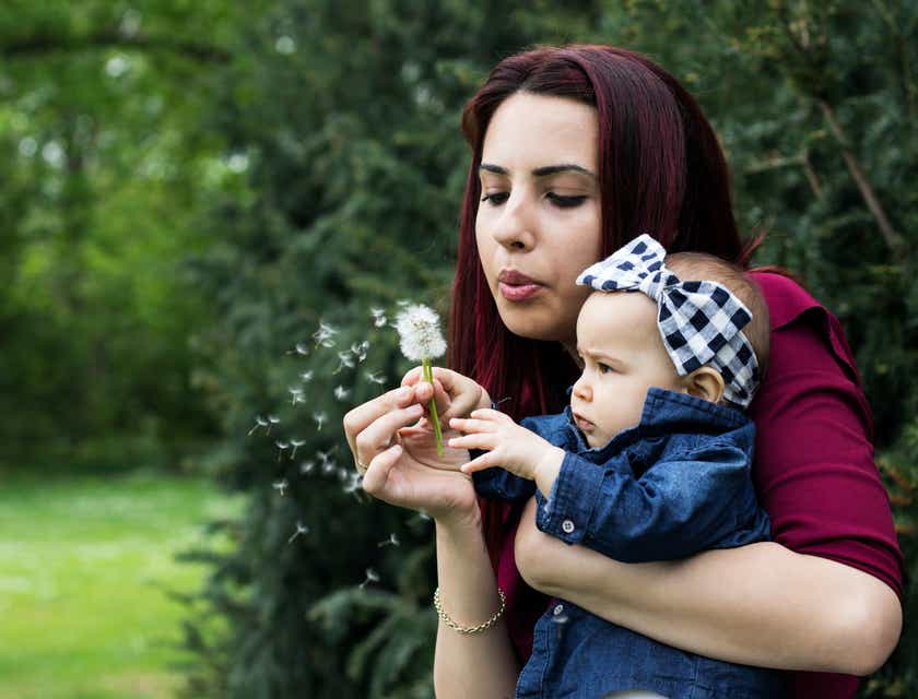A mom holding her baby daughter while blowing a dandelion.