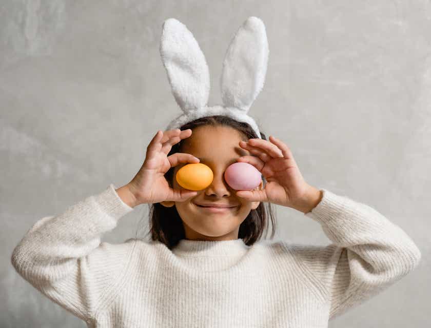 Ein Mädchen hat sich als Hase verkleidet und hält verspielt zwei Eier vor ihre Augen.