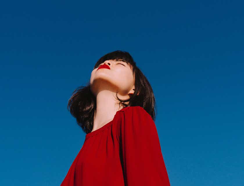 Eine stilvoll im roten Pullover gekleidete Frau steht unter einem wolkenlosen, blauen Himmel und genießt den Sonnenschein.
