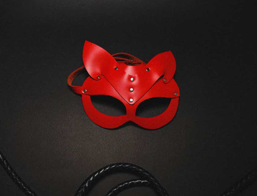 Een foto van een sexy rood masker en een zwarte zweep uit een seksshop tegen een donkere achtergrond.
