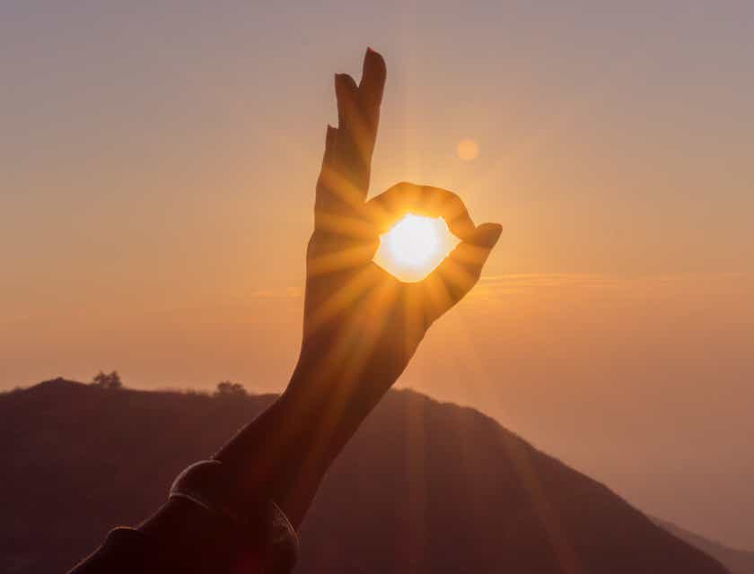Zbliżenie na zachodzące słońce ujęte w środku dłoni wykonującej palcami znak „ok”.
