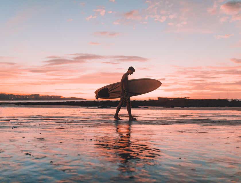 Um surfista andando na areia da praia e segurando uma prancha durante o pôr do sol.